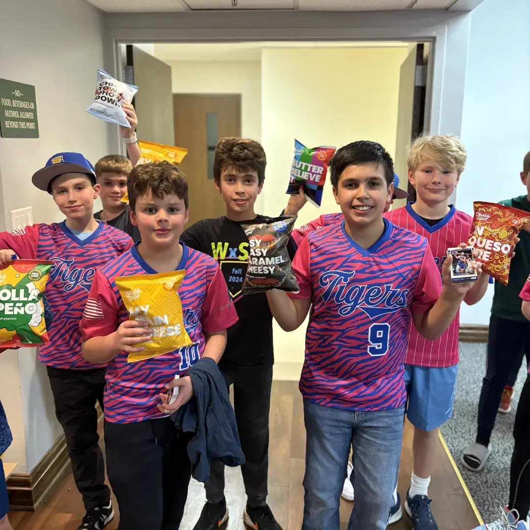 Youth baseball players hold popcorn bags after a fundraising event, smiling in their team jerseys inside a school hallway.