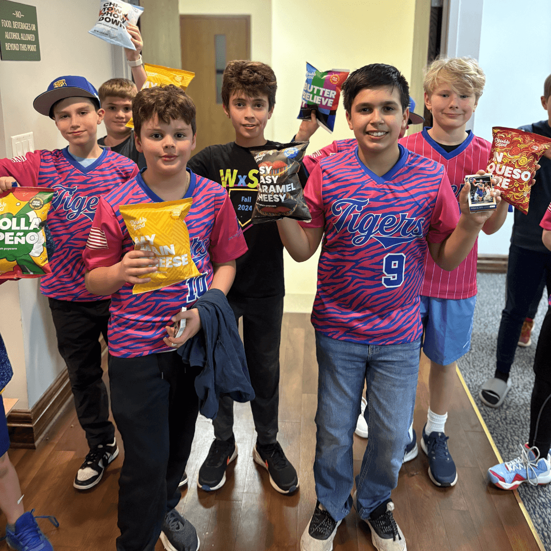 Youth baseball players hold popcorn bags after a fundraising event, smiling in their team jerseys inside a school hallway.