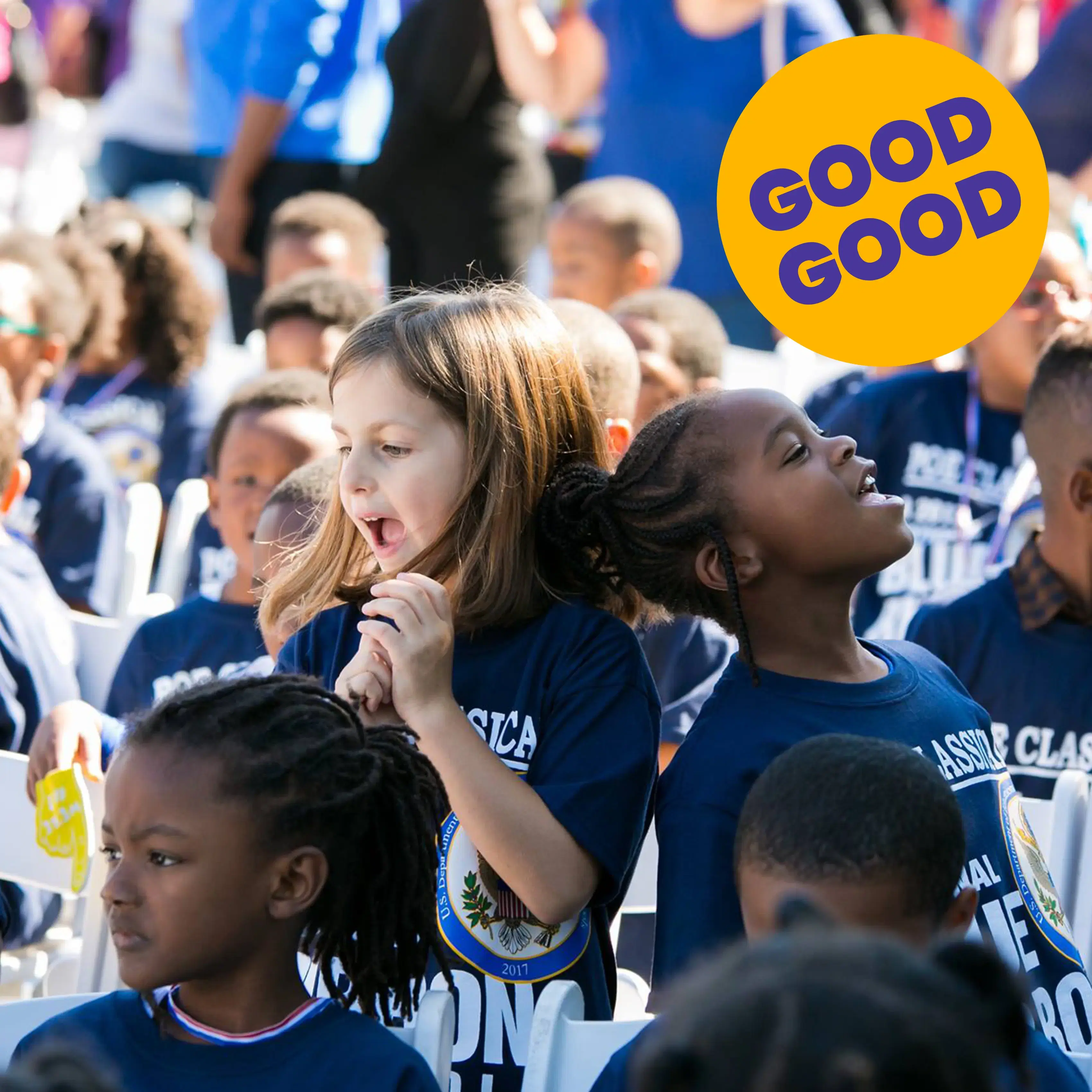 Children wearing matching school shirts cheer at an outdoor event, celebrating a successful school fundraiser with a bright “Good Good” sign overhead.