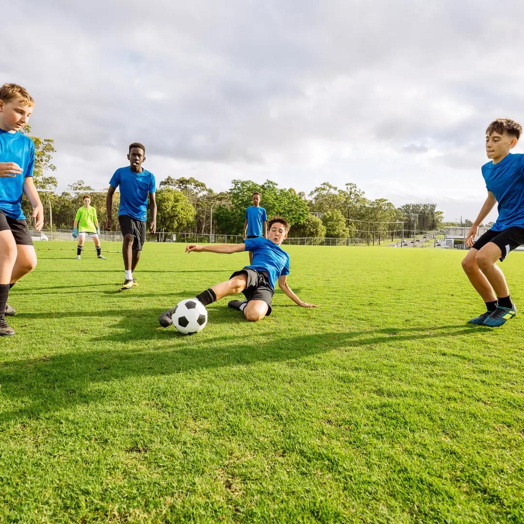 Boys in blue jerseys playing soccer on a green field. One is sliding to kick the ball while others are nearby. Trees and cloudy sky in the background.