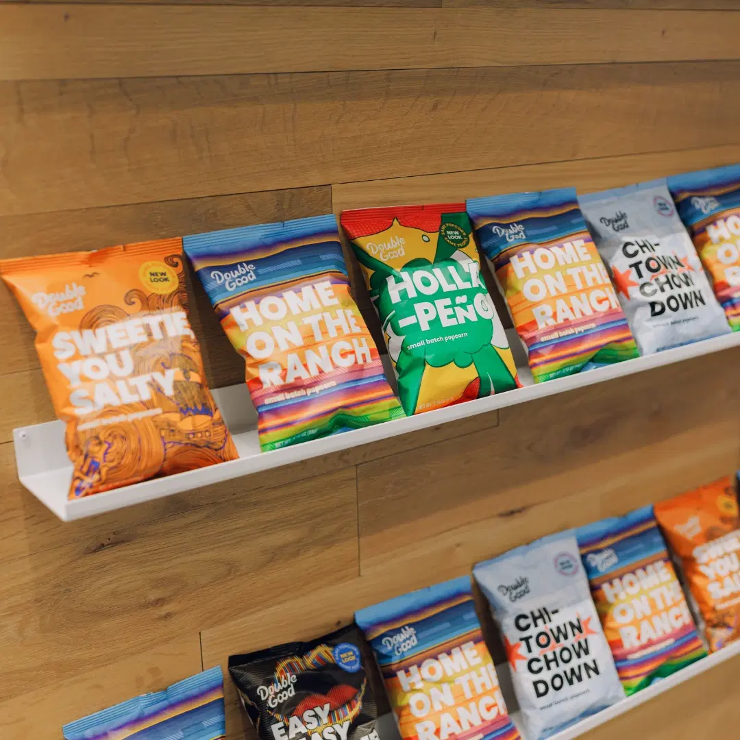 A shelf displaying colorful bags of popcorn with flavors like "Sweetie You Salty," "Home on the Ranch," and "Holla-Peno" against a wooden wall.