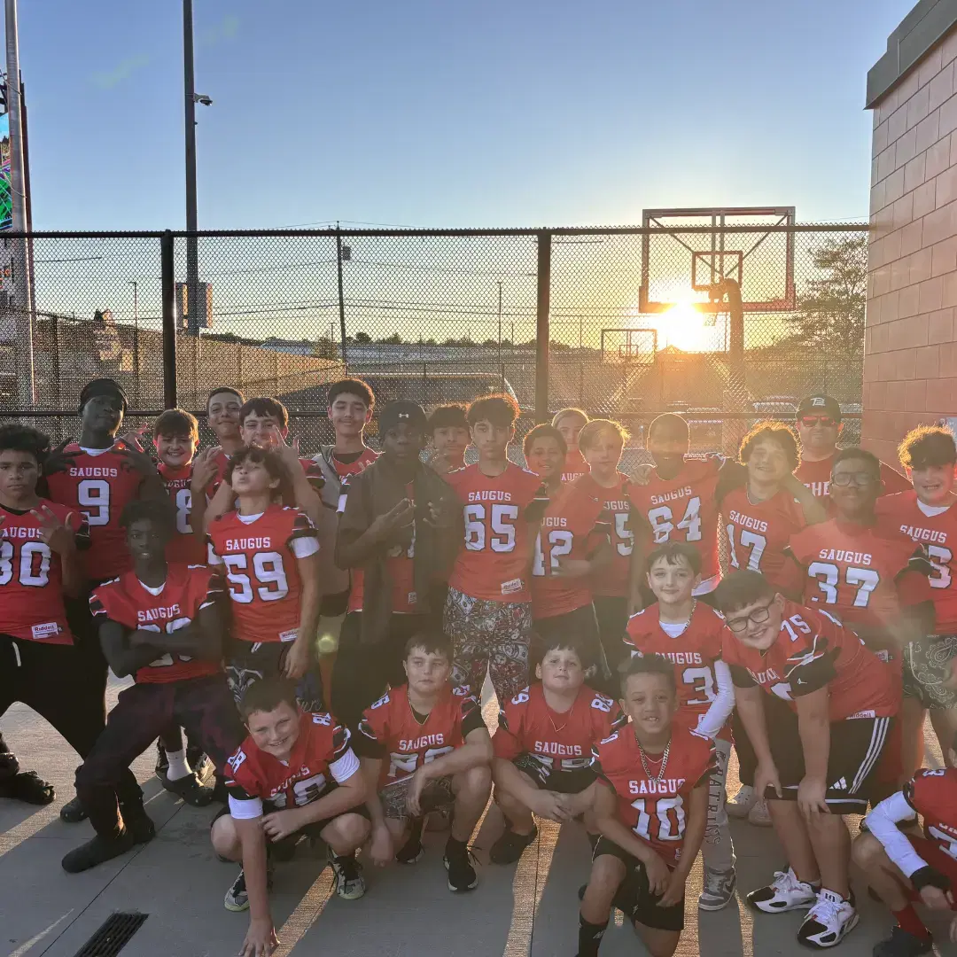 A youth football team wearing red "Saugus" jerseys poses together outside near a basketball court during sunset.