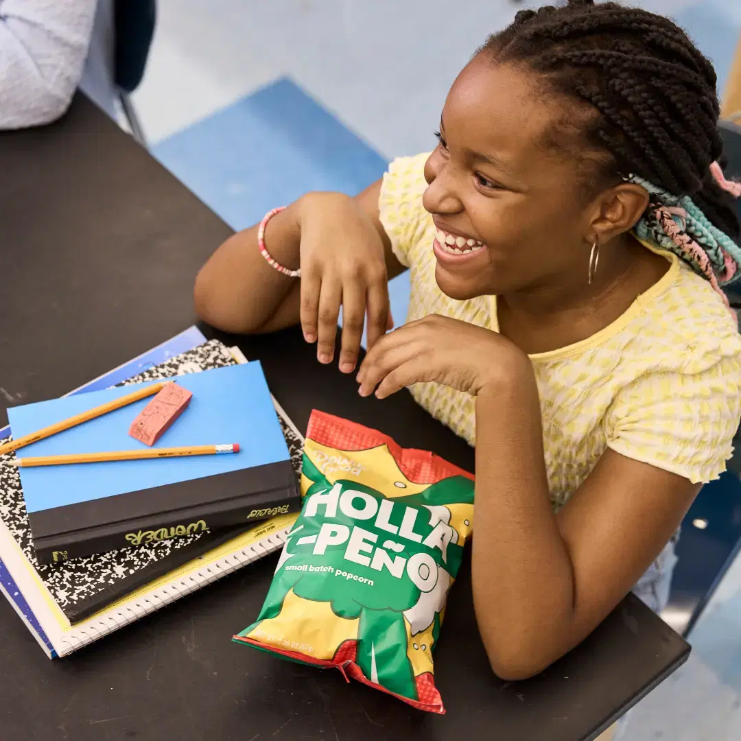 Smiling student at a classroom desk with notebooks, pencils, and a bag of Double Good popcorn, representing the Best Fundraiser option for schools and groups.